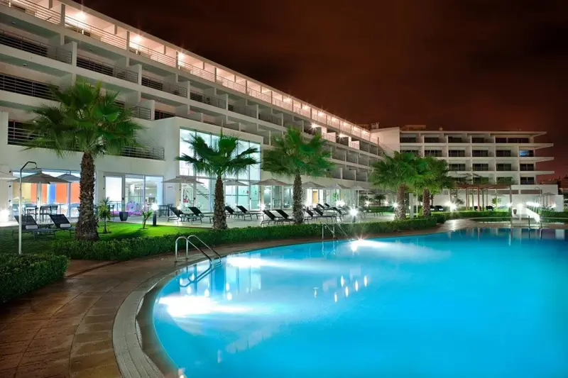 Piscine illuminée au premier plan d'un bâtiment moderne la nuit.