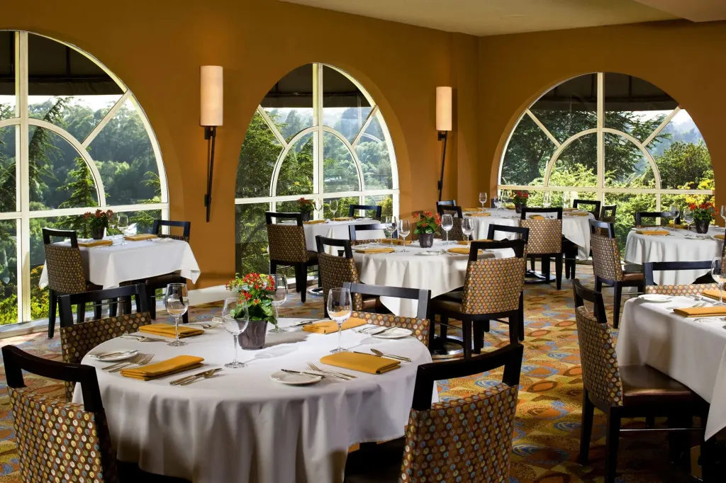 Une salle lumineuse avec des tables dressées pour un repas face à un paysage verdoyant.