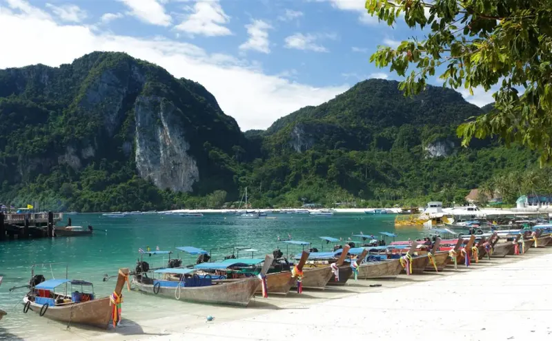 Des bateaux colorés sont amarrés sur une plage de sable blanc bordée de montagnes verdoyantes.