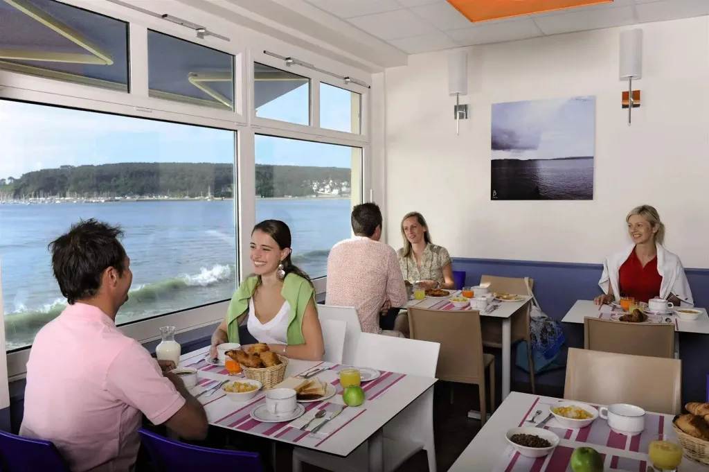 Une salle lumineuse avec des tables où des clients prennent leur petit-déjeuner face à une vue sur la mer.