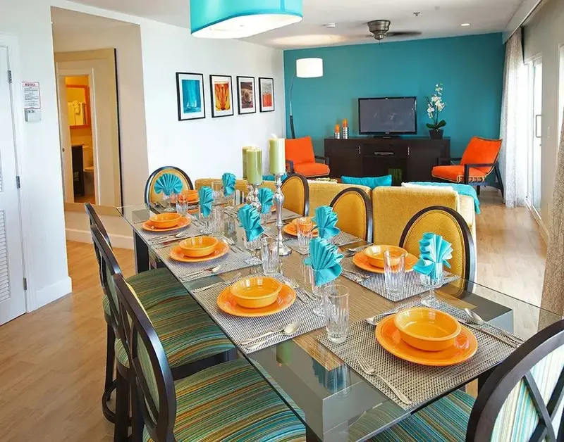 Une grande table à manger est dressée pour un repas avec des couverts et de la vaisselle colorée.