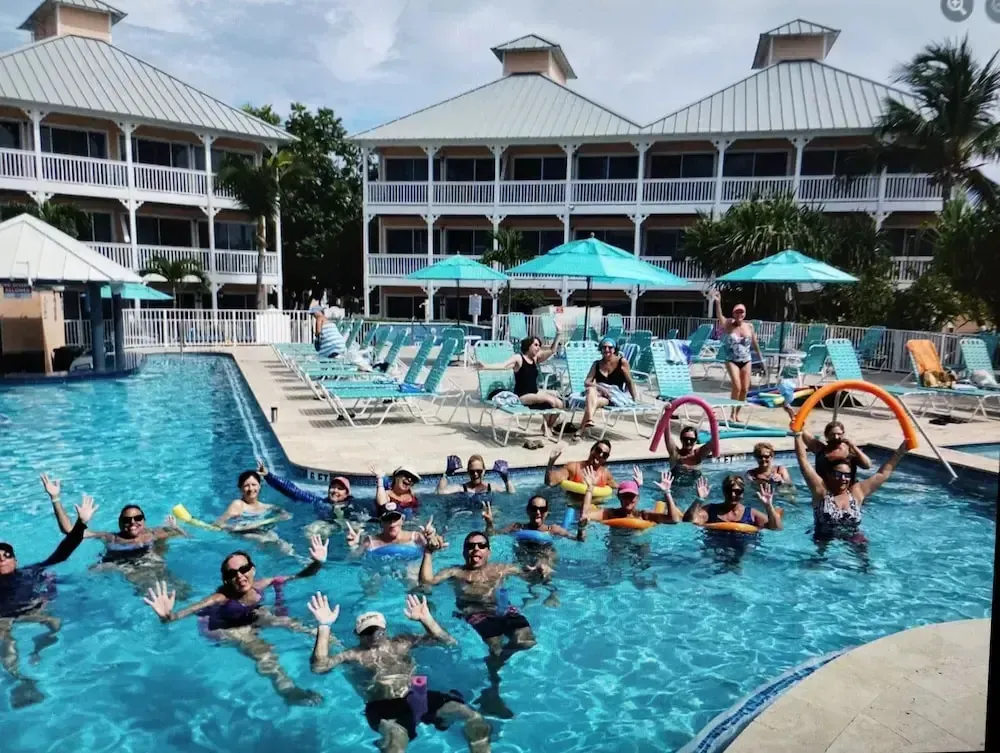 Personnes souriantes dans une piscine ensoleillée, entourées de transats et de parasols.