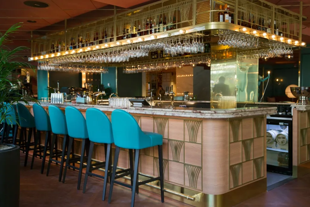 Un long comptoir de bar avec des tabourets turquoise et des rangées de verres.