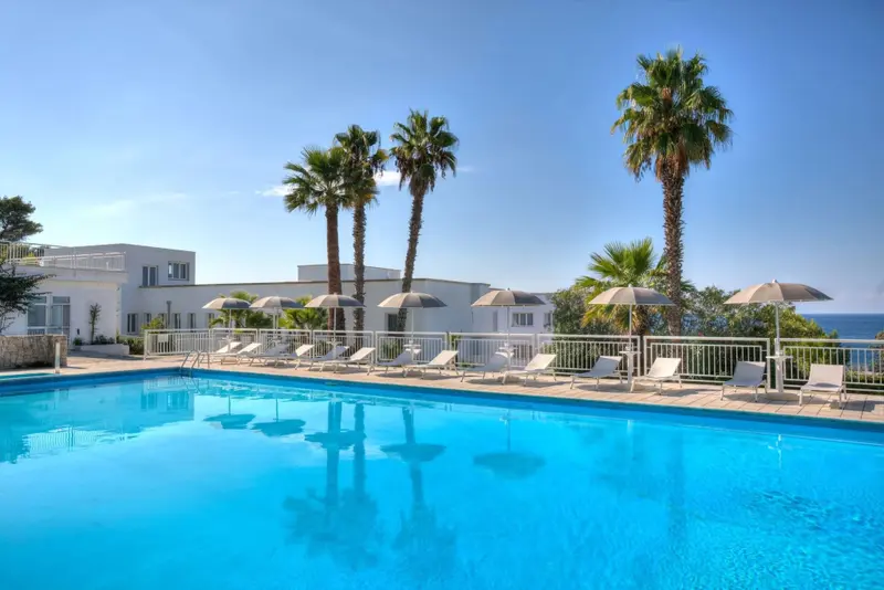 Une piscine turquoise bordée de transats sous des palmiers, avec un bâtiment blanc en arrière-plan.