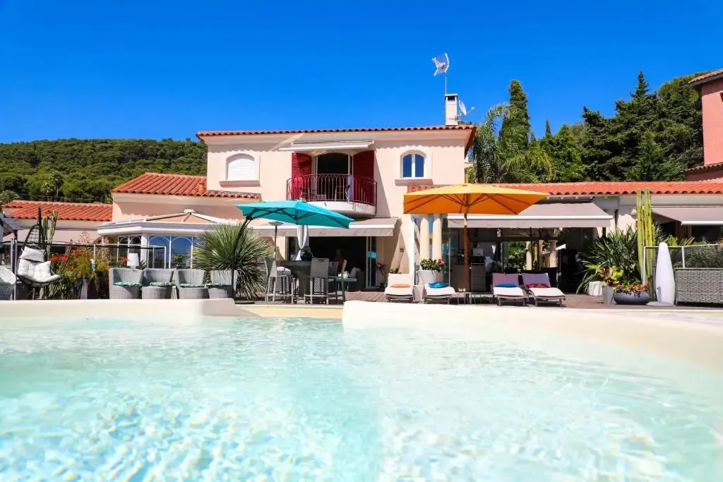 Une piscine turquoise bordée d'une terrasse avec des transats et des parasols devant un bâtiment rose.