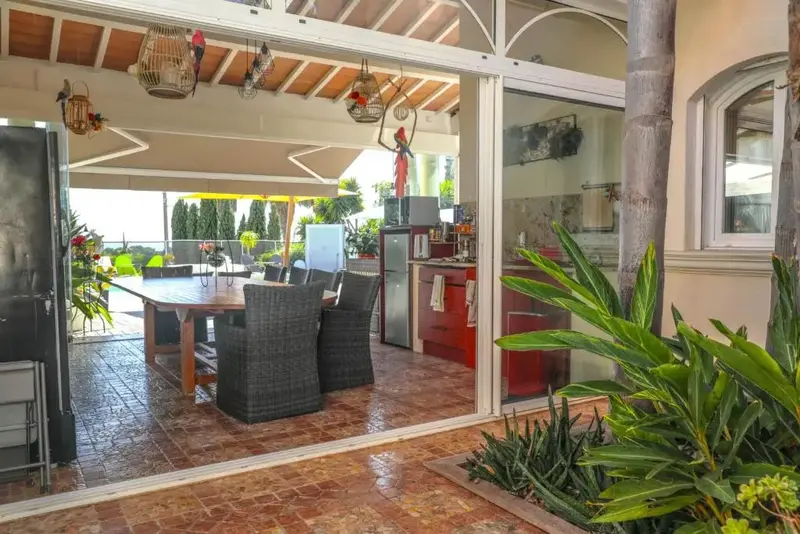 Une grande table à manger en bois est entourée de chaises sur une terrasse carrelée.
