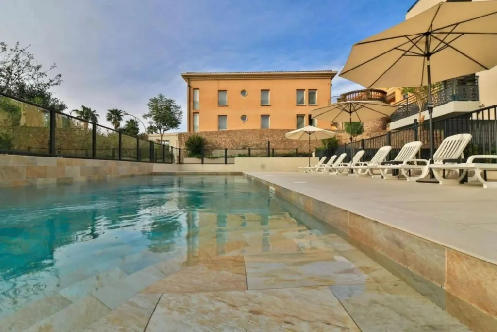 Une piscine rectangulaire avec des transats et parasols, devant un bâtiment à l'architecture classique.