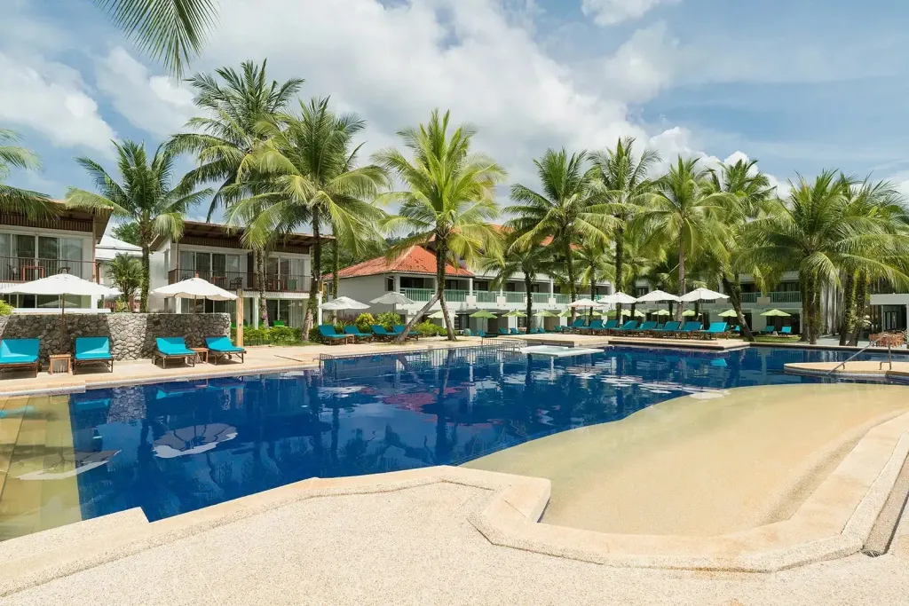 Une piscine bordée de transats et de palmiers devant des bâtiments.