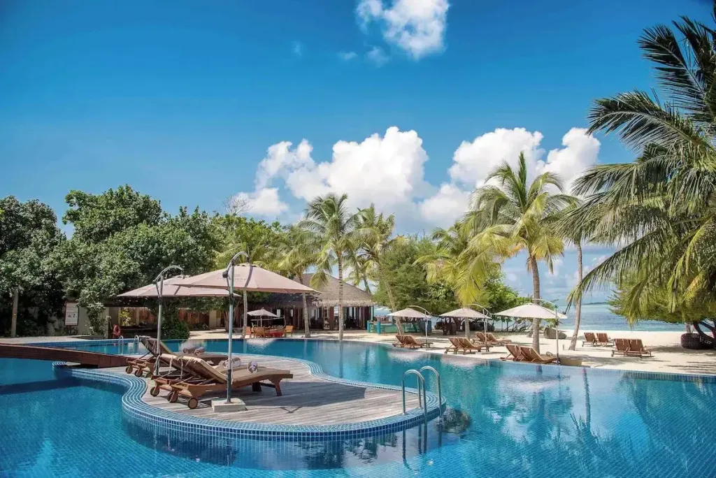 Une piscine bleue bordée de transats sous des parasols, avec une plage de sable fin en arrière-plan.