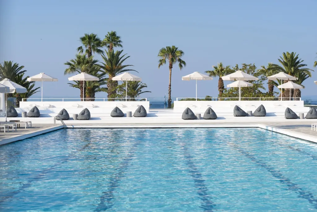 Une piscine rectangulaire bordée de transats et de parasols blancs, avec des palmiers en arrière-plan et la mer au loin.