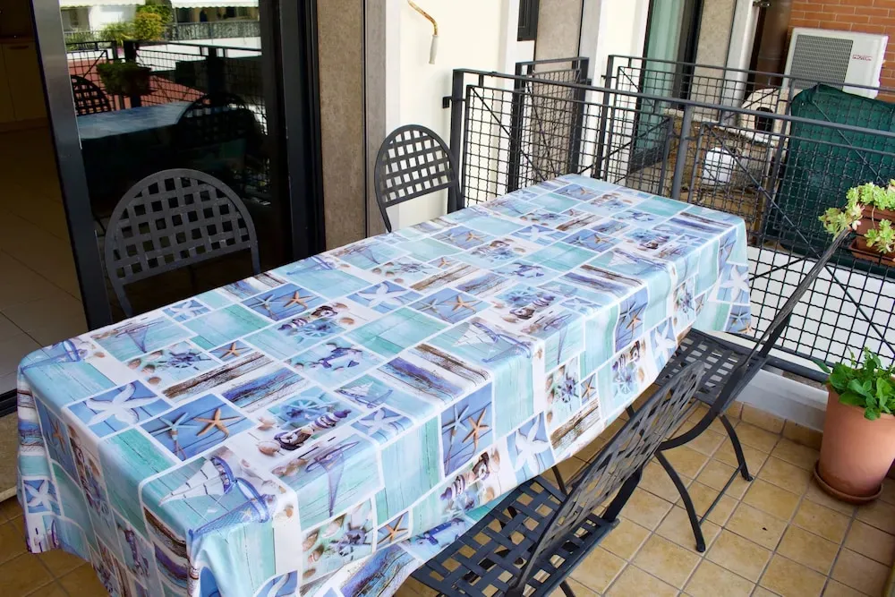 Une table recouverte d'une nappe à motifs marins est entourée de chaises sur une terrasse carrelée.