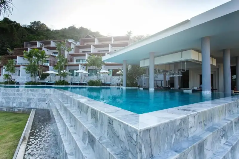 Une piscine bleue bordée de marbre blanc offre une vue sur un bâtiment moderne et une végétation luxuriante.