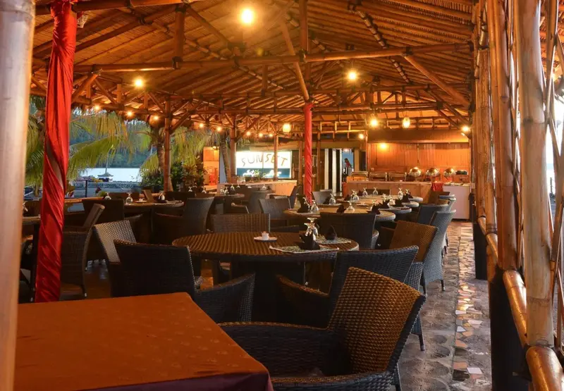 Une salle à manger extérieure abritée sous une toiture en bois et bambou, avec des tables et des chaises en osier.