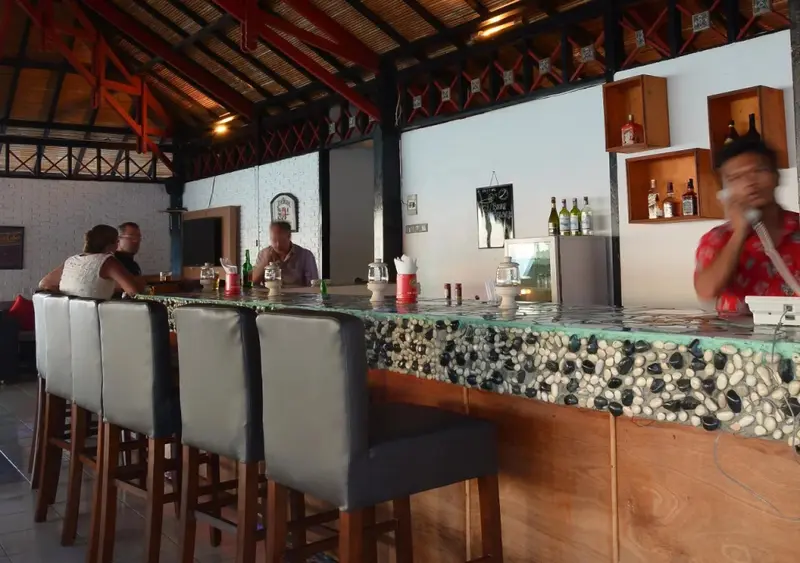 Un comptoir de bar est desservi par un employé, des clients sont assis sur des tabourets.