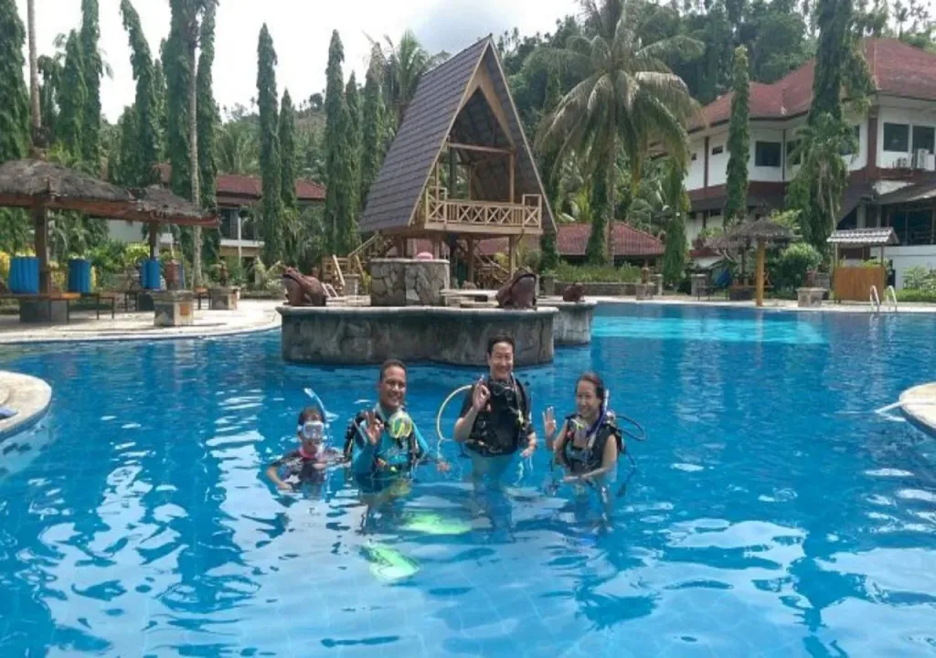 Des personnes équipées se tiennent dans une piscine ensoleillée devant un bâtiment à l'architecture distinctive.