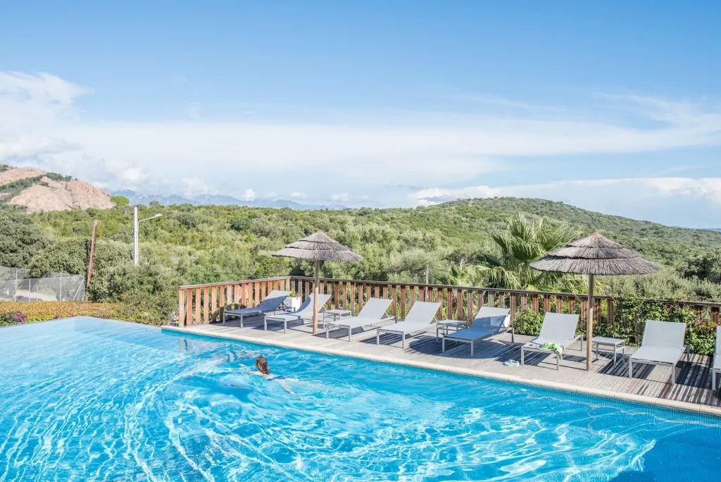 Une piscine rectangulaire bordée de transats sous des parasols, avec une vue sur une colline verdoyante.