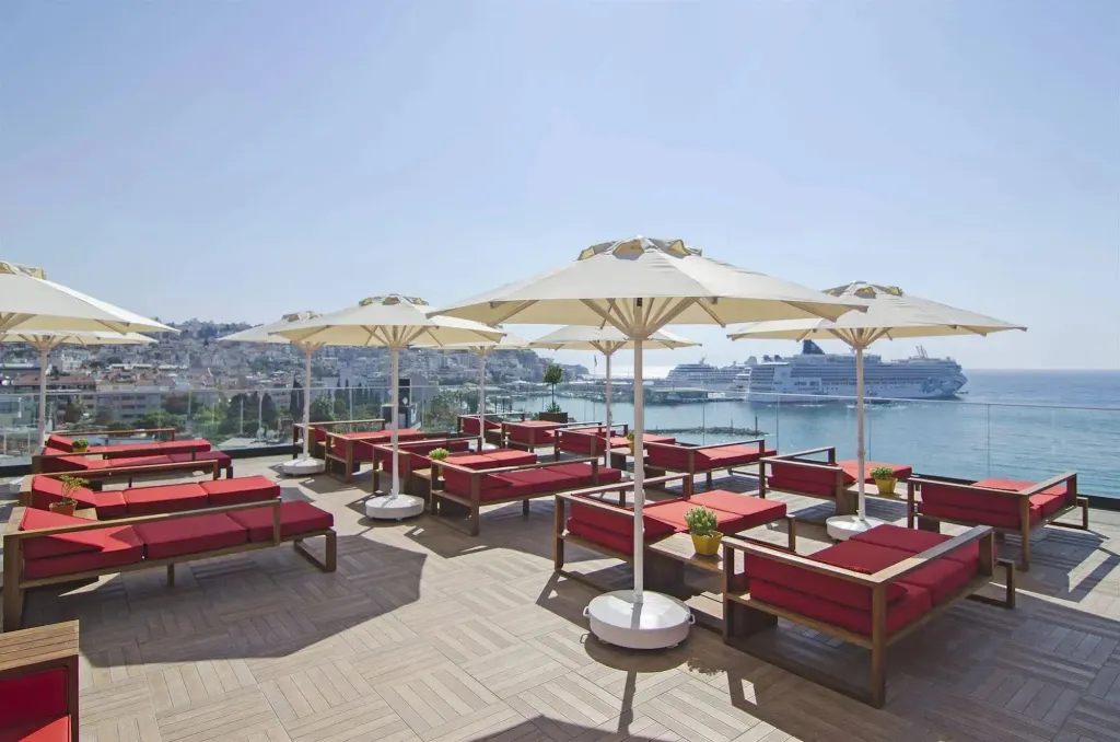 Terrasse aménagée avec des transats rouges et des parasols blancs offrant une vue sur la mer et la ville.