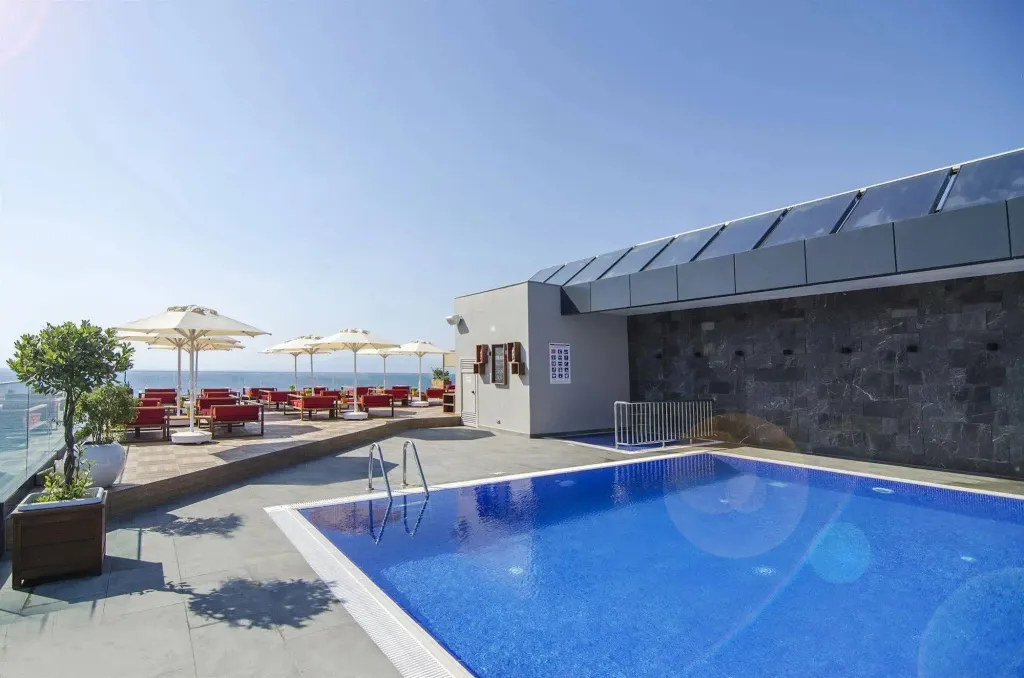 Une piscine bleue avec des transats rouges et des parasols blancs sur une terrasse ensoleillée.