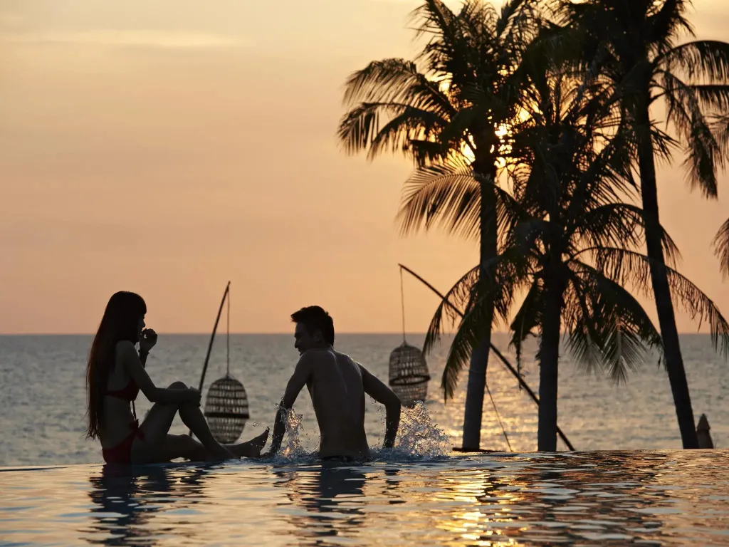 Deux personnes se détendent au bord d'une piscine face à la mer au coucher du soleil.