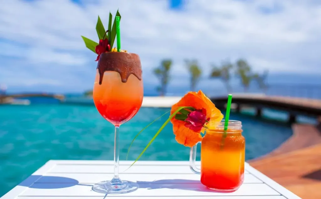 Deux cocktails colorés sont présentés sur une table blanche près d'une piscine turquoise.