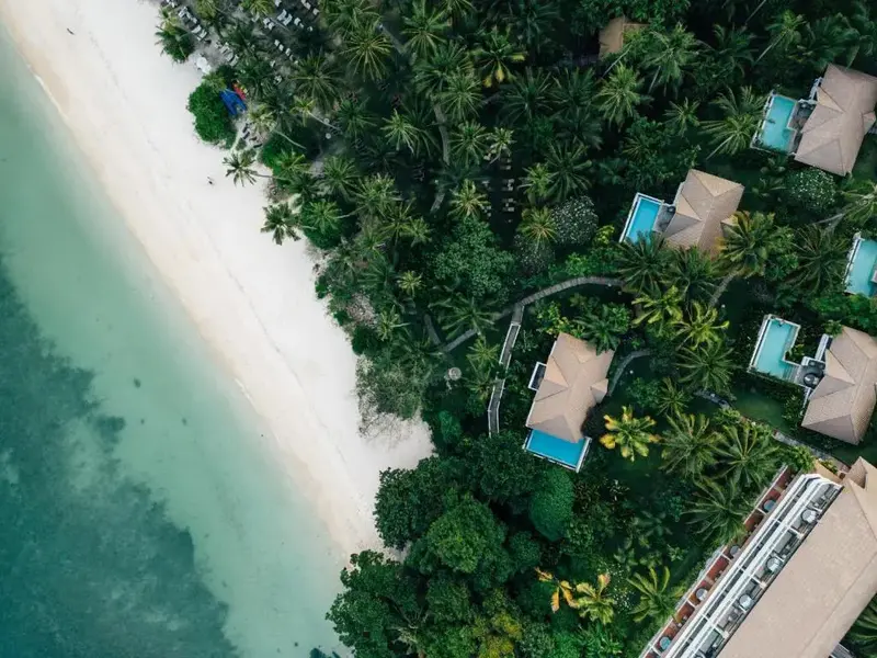Vue aérienne d'une plage bordée de végétation luxuriante et de bâtiments aux toits de chaume.