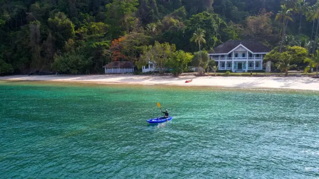 Une personne navigue en kayak sur une eau turquoise devant une plage de sable fin et une végétation luxuriante.
