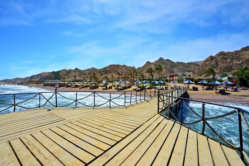 Un ponton en bois mène vers une plage bordée de parasols et de chaises longues.