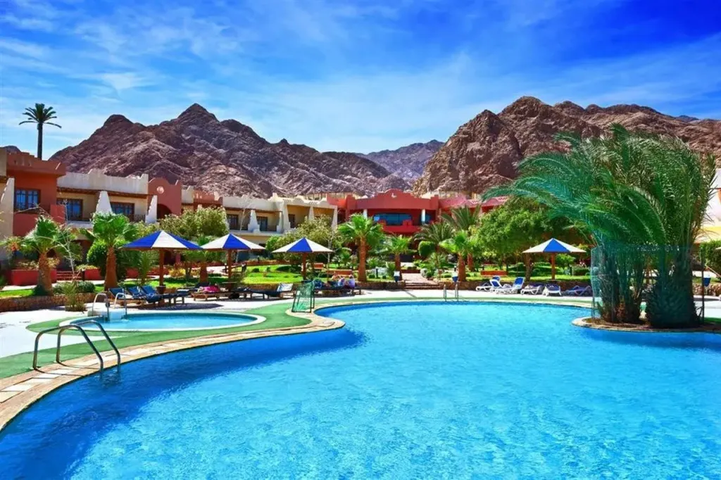 Une piscine turquoise bordée de transats et de palmiers, avec des bâtiments aux tons chauds en arrière-plan.