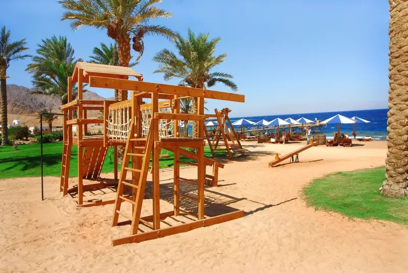 Structure de jeux en bois sur une plage de sable fin, avec des transats et des parasols en arrière-plan.