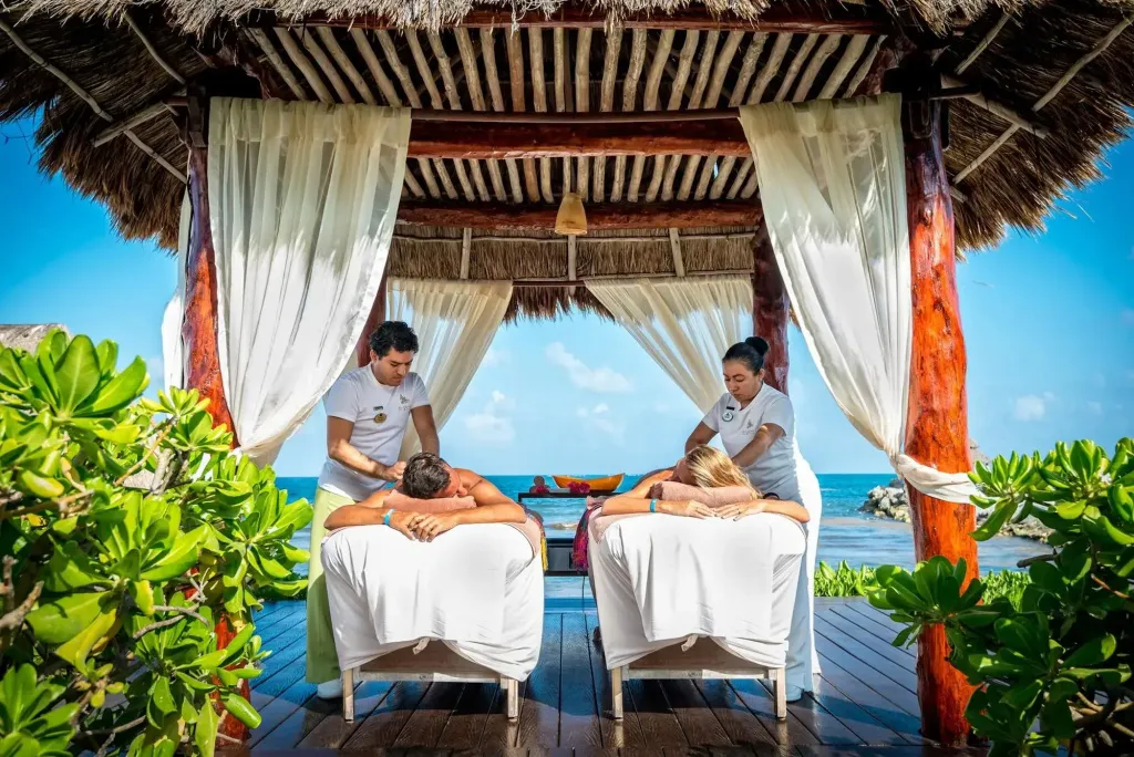 Deux personnes allongées reçoivent un massage dans un espace extérieur ombragé face à la mer.