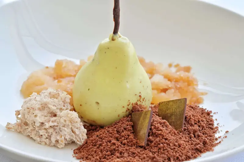 Une poire pochée est présentée avec des morceaux d'ananas, une mousse et une poudre de cacao sur une assiette blanche.