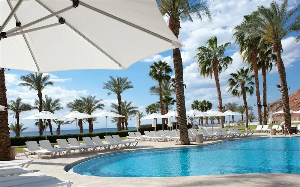 Une piscine bordée de transats blancs sous des parasols, avec des palmiers et la mer en arrière-plan.