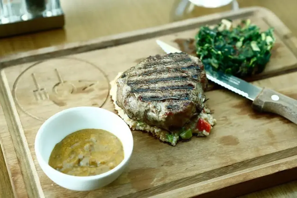 Une portion de viande grillée est servie avec une sauce et des légumes verts sur une planche en bois.