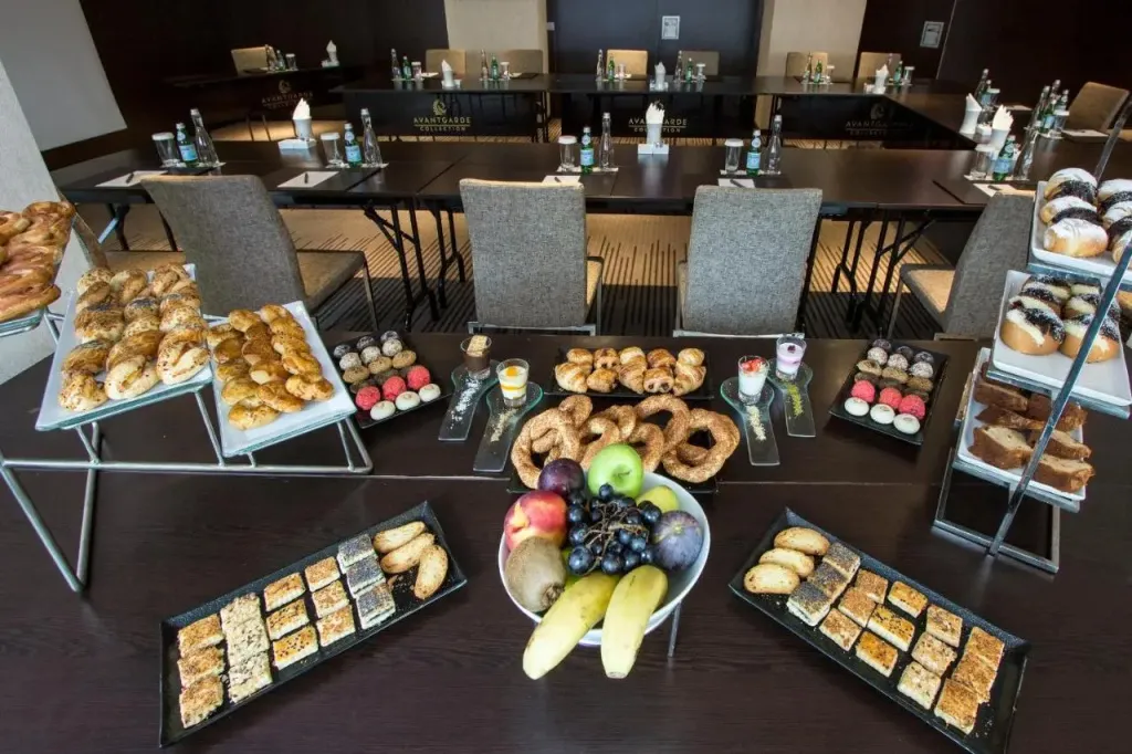 Une longue table est dressée de pâtisseries, de fruits et de boissons dans une salle de réunion.