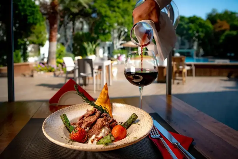 Une assiette de viande accompagnée de légumes est servie avec du vin rouge sur une table en extérieur.
