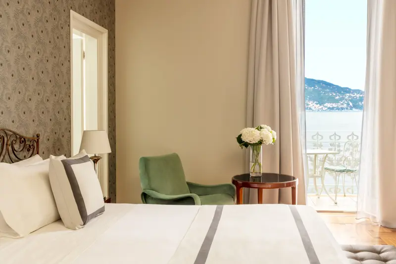 Une chambre lumineuse avec un lit soigné et un fauteuil vert près d'une fenêtre ouverte sur la mer.