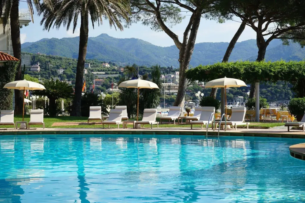 Une piscine turquoise bordée de transats sous des parasols, avec une vue sur la mer et les montagnes.