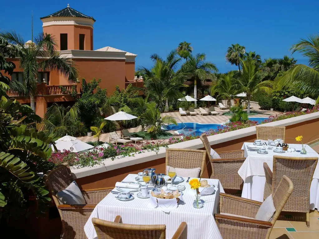Tables dressées pour le petit-déjeuner sur une terrasse ensoleillée avec vue sur une piscine et des bâtiments.