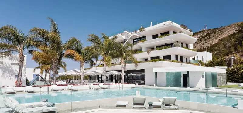 Vue d'un bâtiment blanc moderne avec une piscine ensoleillée et des palmiers.