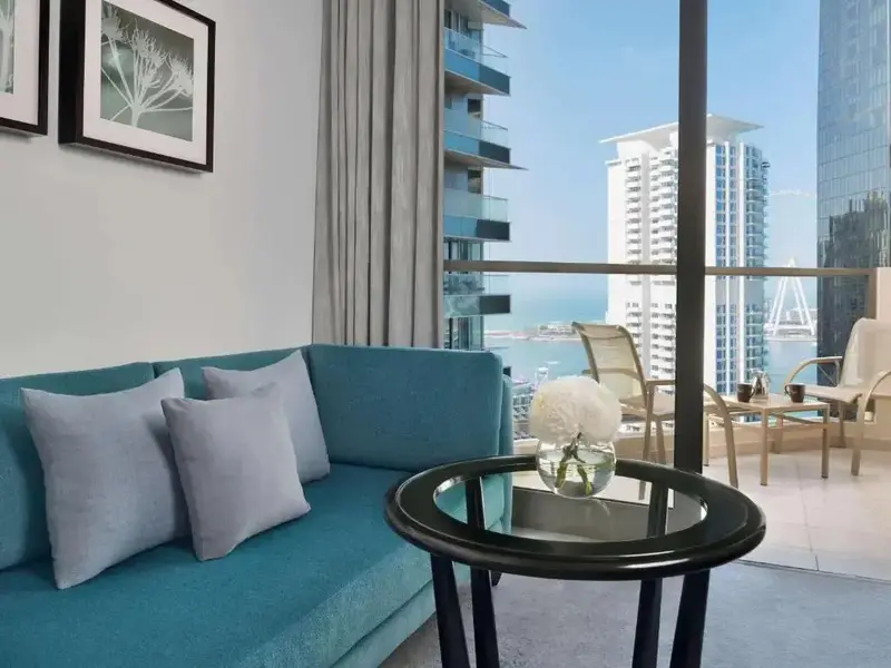 Un canapé bleu turquoise est placé devant une grande baie vitrée donnant sur la mer et les buildings.