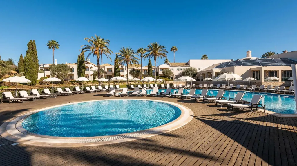 Une piscine rectangulaire entourée de transats sous des parasols, avec des bâtiments en arrière-plan.