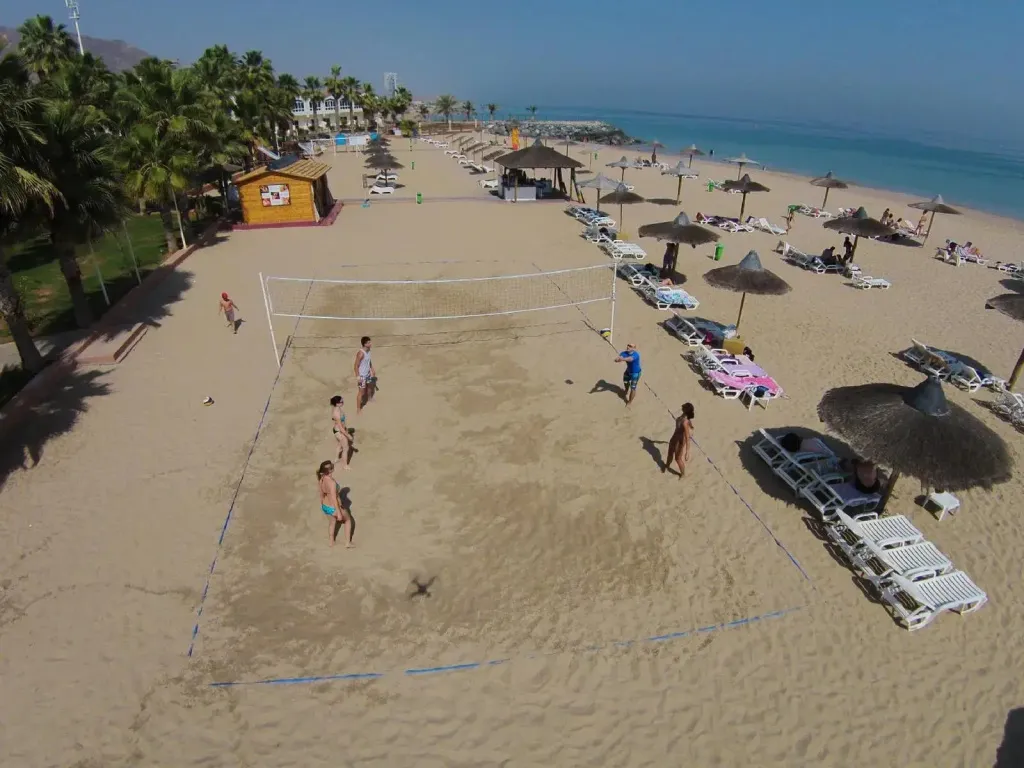 Une plage de sable fin avec des personnes jouant au volleyball et des transats à l'ombre de parasols.