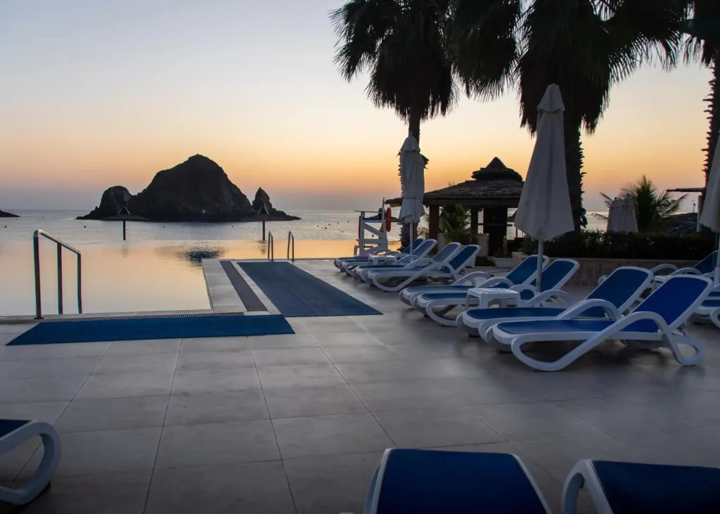 Transats bleus alignés au bord d'une piscine face à la mer au coucher du soleil.
