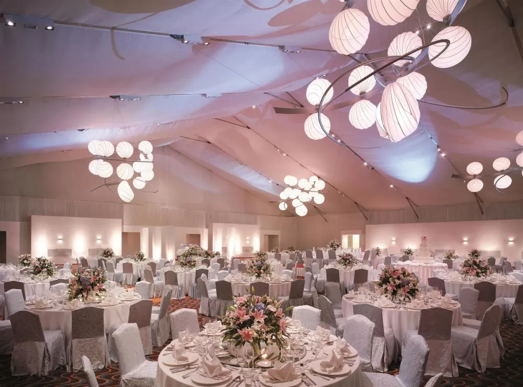 Une grande salle lumineuse est aménagée pour un banquet avec des tables rondes nappées et des chaises.