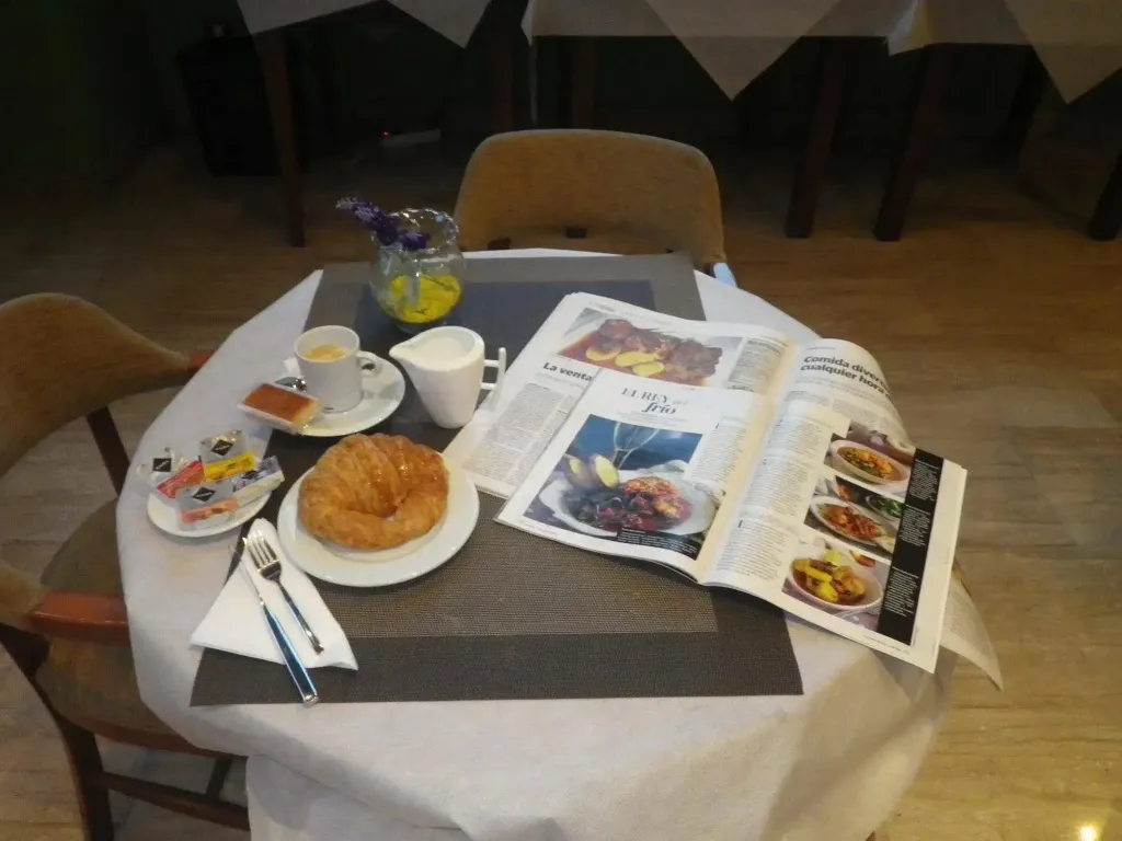 Une table est dressée pour le petit-déjeuner avec une viennoiserie, une boisson chaude et un journal.