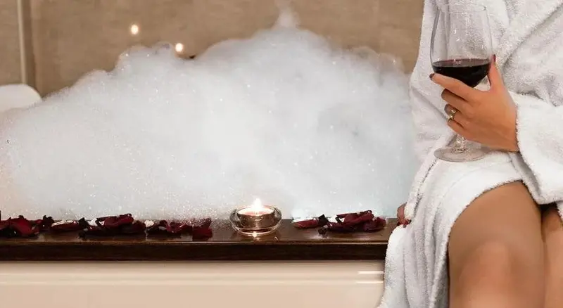 Une personne en peignoir se détend dans un bain moussant avec un verre de vin et une bougie.