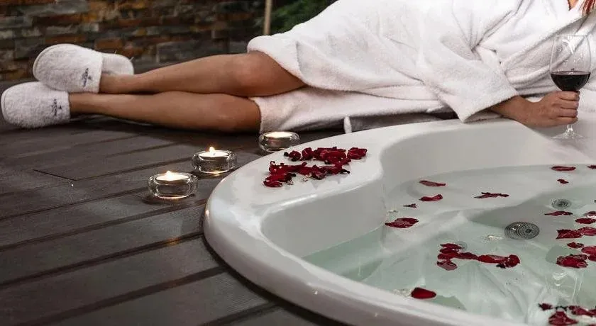 Une personne se détend dans un jacuzzi extérieur décoré de pétales de roses, un verre de vin à la main.