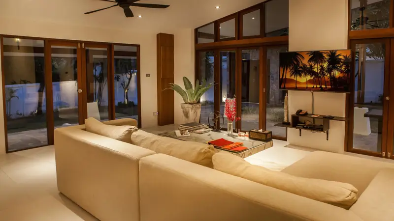 Un salon lumineux avec canapés beiges et grandes baies vitrées donnant sur un jardin tropical.