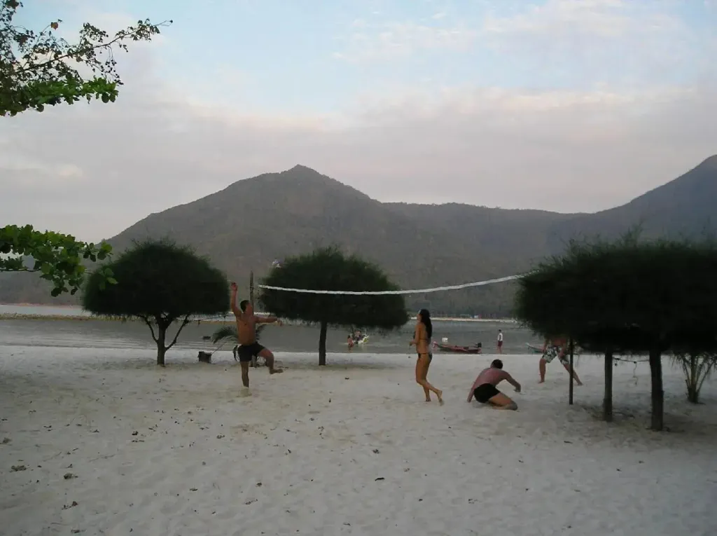 Des personnes jouent au volleyball sur une plage de sable fin avec des arbres et des montagnes en arrière-plan.