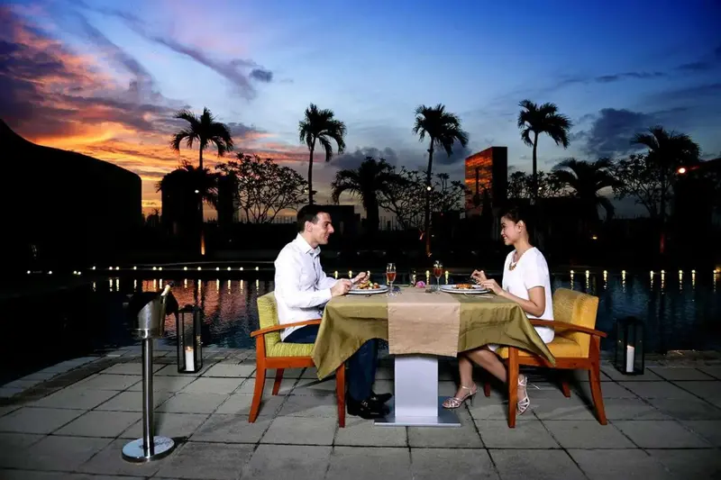Un couple dîne à une table en extérieur au bord d'une piscine illuminée au coucher du soleil.